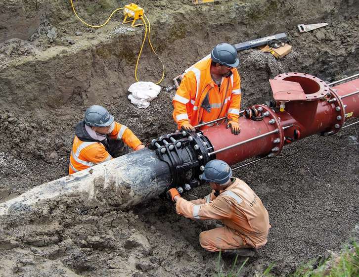 Three utility workers wearing hard hats and hi-vis clothing securing a large pipe joint in an excavation trench