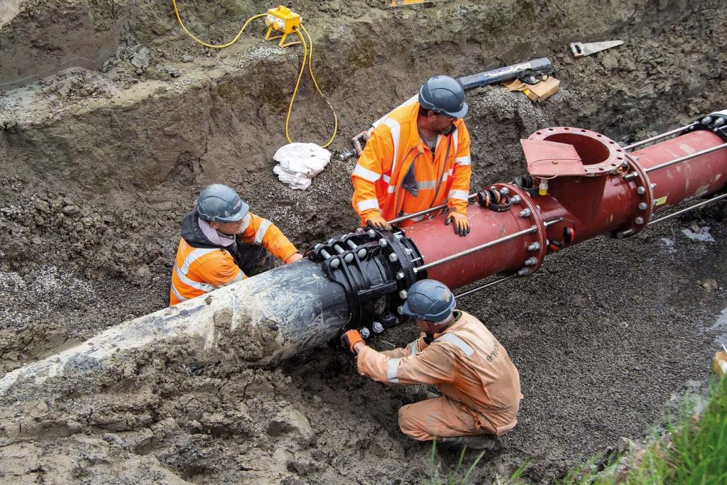 Three utility workers wearing hard hats and hi-vis clothing connecting large pipes in a trench