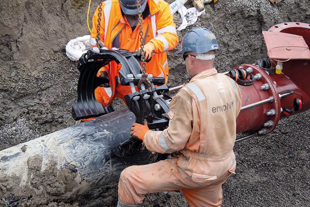 Two utility workers fitting a heavy clamp onto a large pipe in a trench