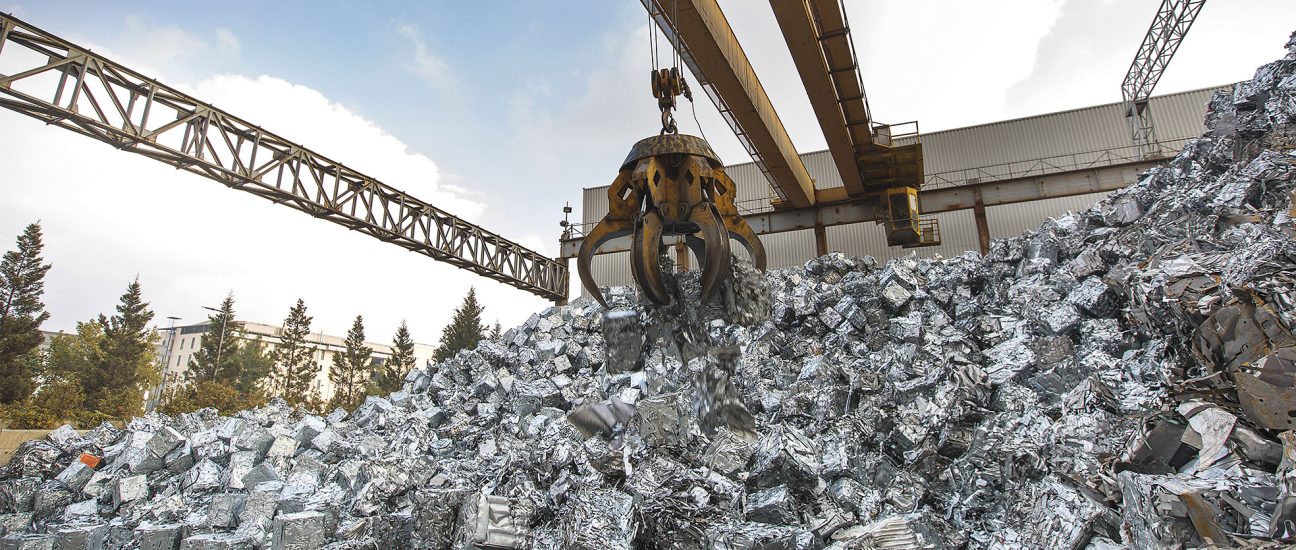 An overhead crane claw lifting crushed metal bales from a large pile of scrap metal