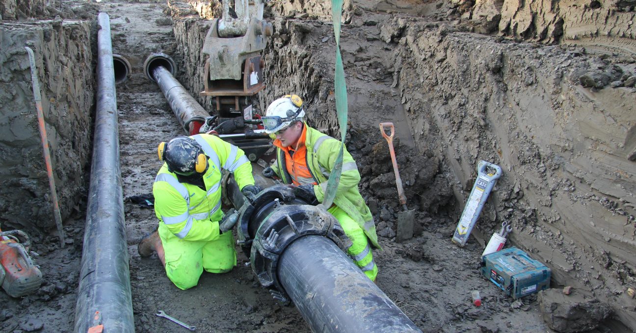 Two workers in high-visibility safety clothing fitting a joint onto a large black pipe in a muddy trench