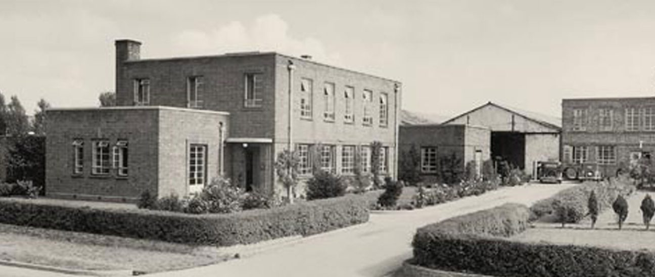 Historic two-storey brick factory building surrounded by manicured hedges and a driveway