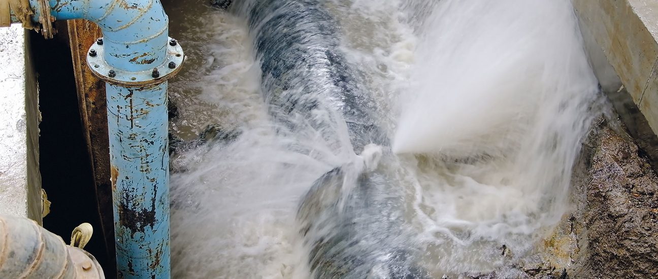 Water spraying forcefully from a burst underground pipe beside a vertical blue pipe