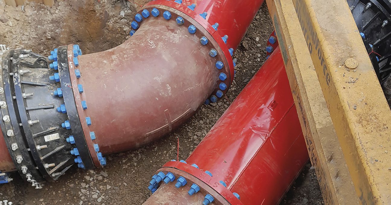 Large red underground pipes connected by flanged joints with blue bolts in a trench