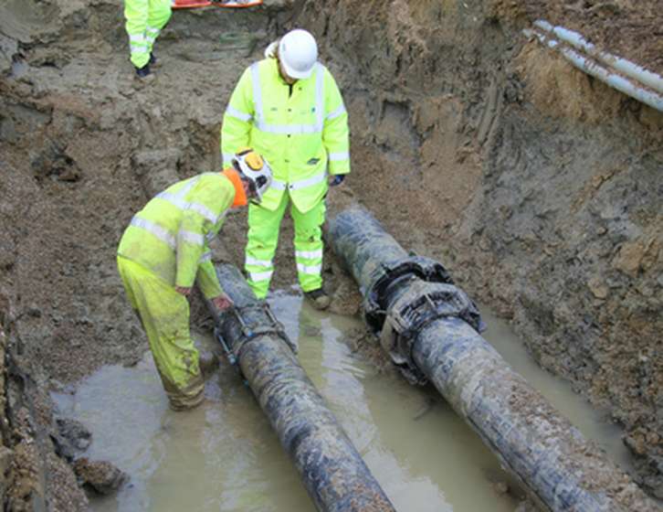 Two utility workers in high-visibility gear inspect large underground pipes in a muddy trench