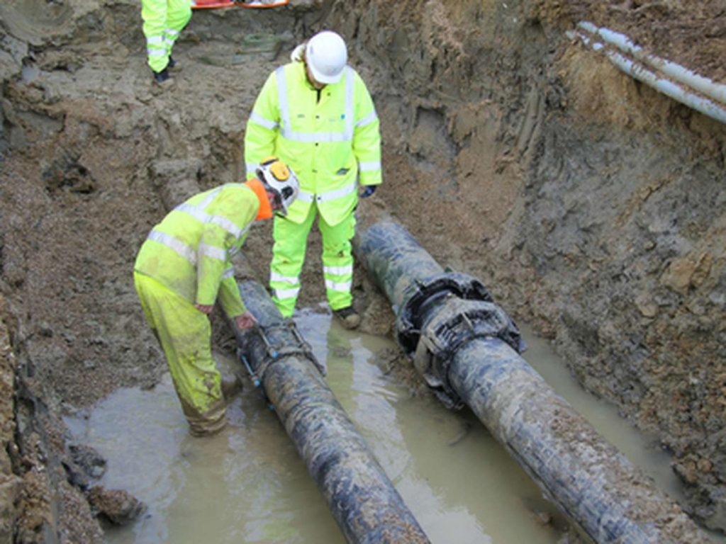 Two workers in high-visibility gear inspecting large underground pipes in a muddy trench
