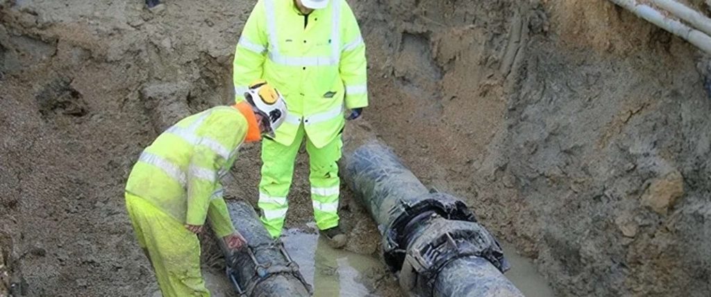 Two utility workers in high-visibility clothing inspecting underground pipes in an excavated trench