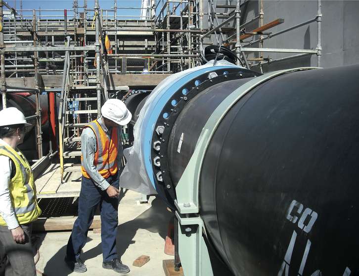 Two workers in hard hats and high-visibility vests inspect a large industrial pipe at a construction site