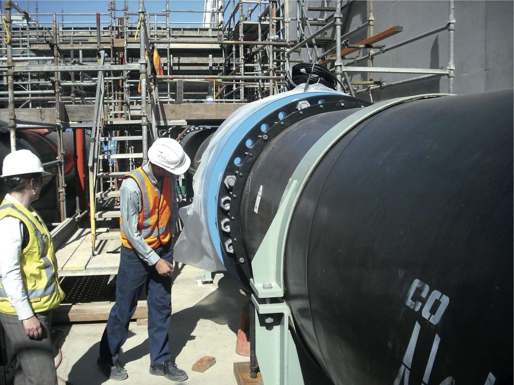 Two workers in hard hats and high-visibility vests inspecting a large industrial pipe on a construction site