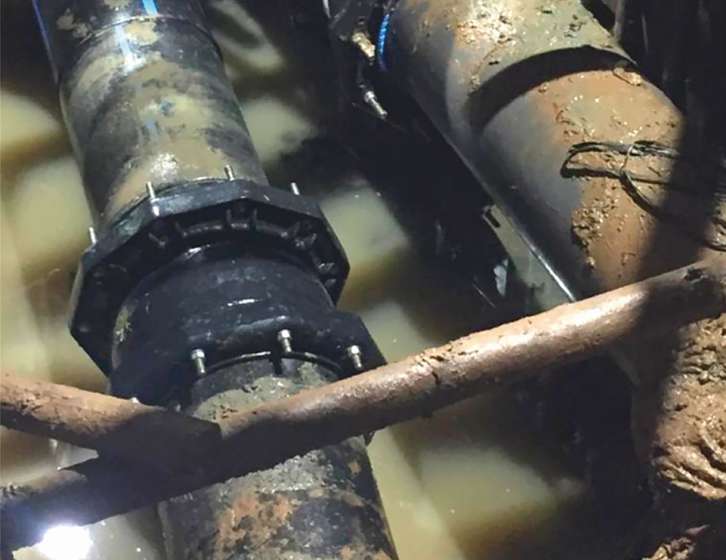 Large black pipes with bolted couplings joined together inside a muddy, water-filled trench