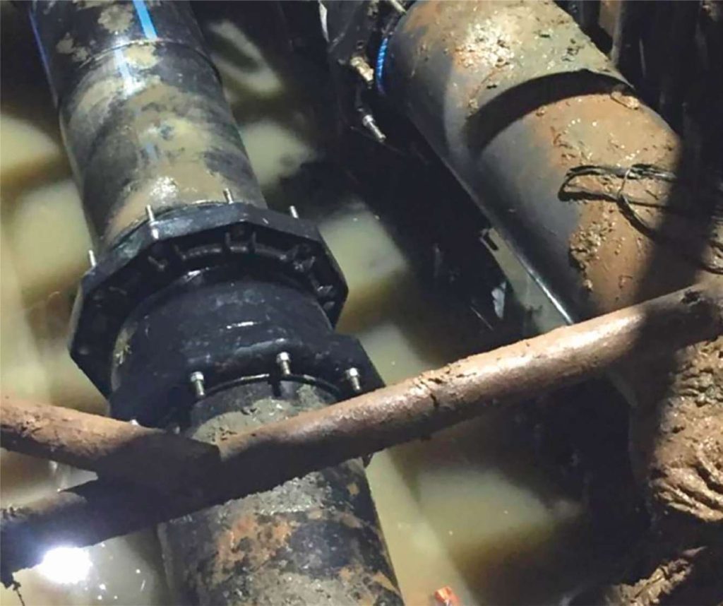 Black pipes connected with a bolted flange coupling in a muddy, water-filled underground trench