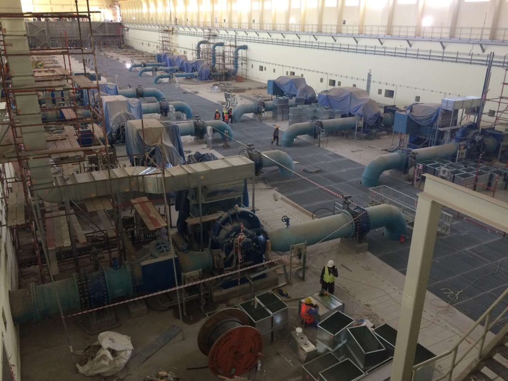 Workers inside a large industrial water pumping station under construction, with large blue pipes and scaffolding