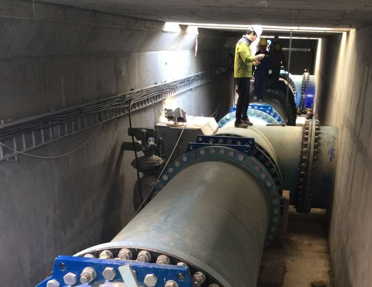 Worker standing on a large industrial pipe inside an underground concrete tunnel