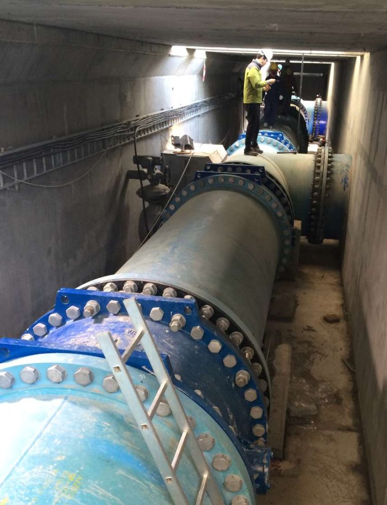 Worker inspecting large blue industrial pipes inside a concrete underground tunnel