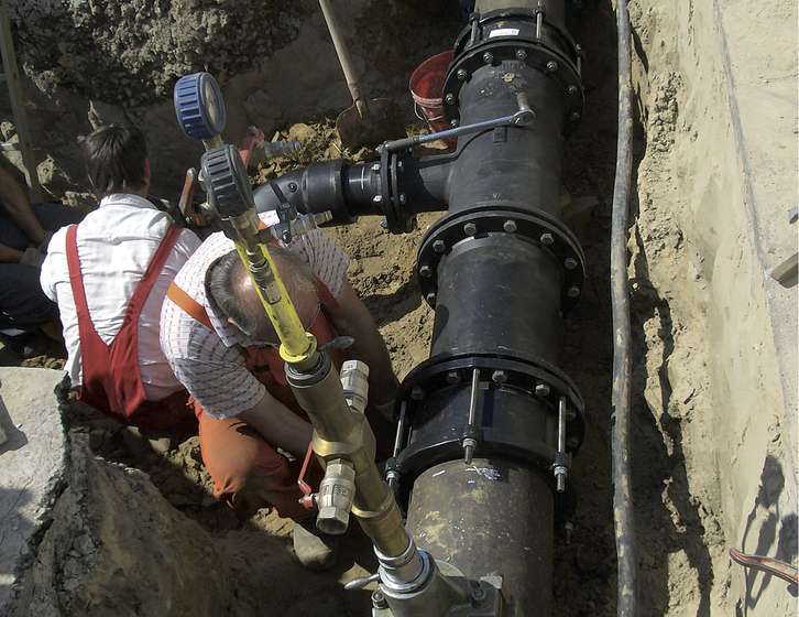 Two workers in a trench installing and testing large black underground pipes fitted with heavy flanges