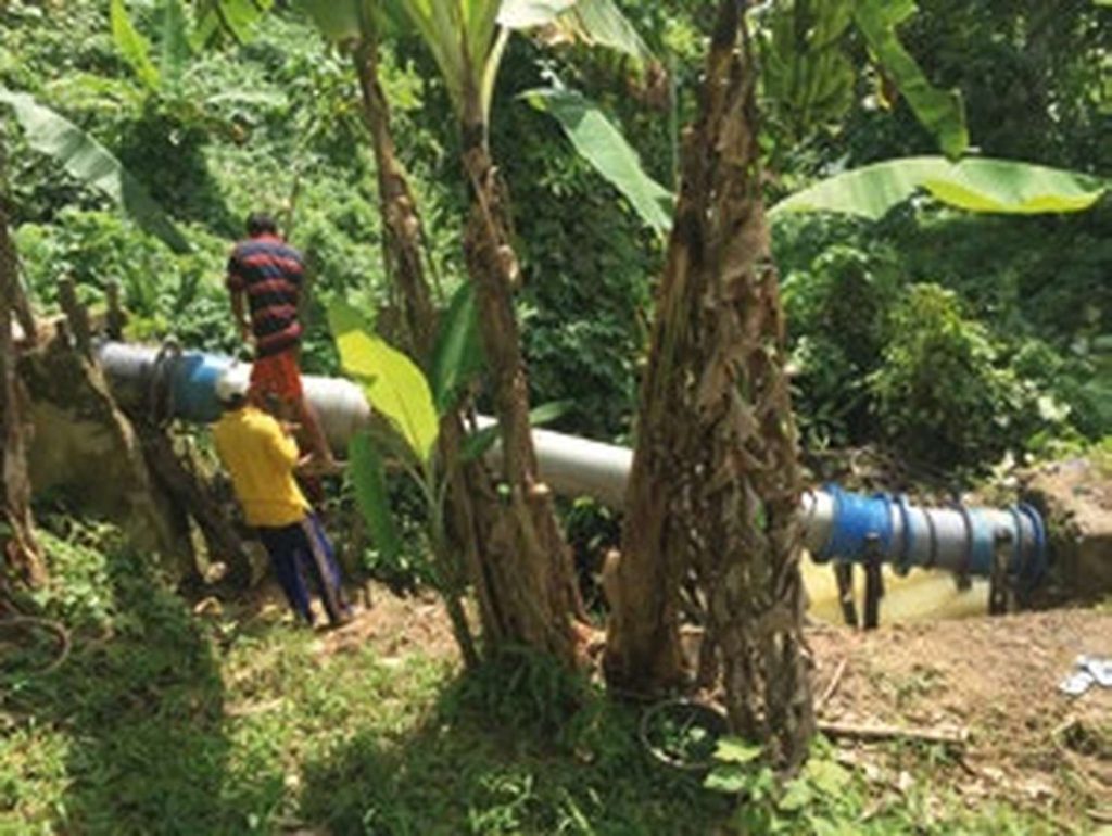 Two workers installing or repairing a large pipe running through lush tropical vegetation