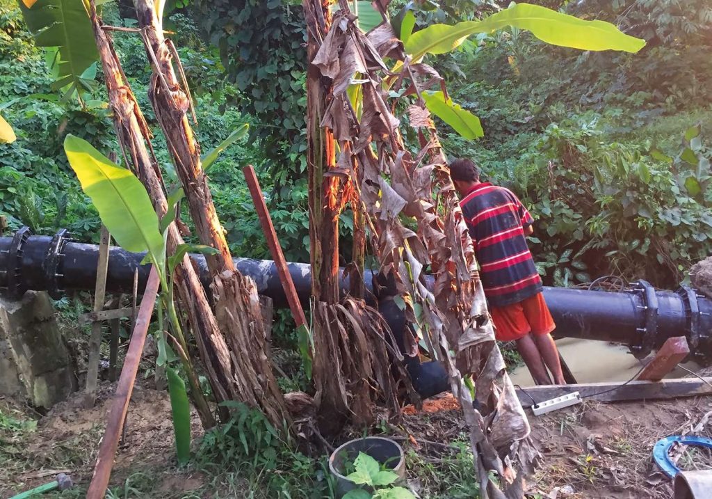 A man standing in a ditch next to a large black pipeline surrounded by dense tropical foliage