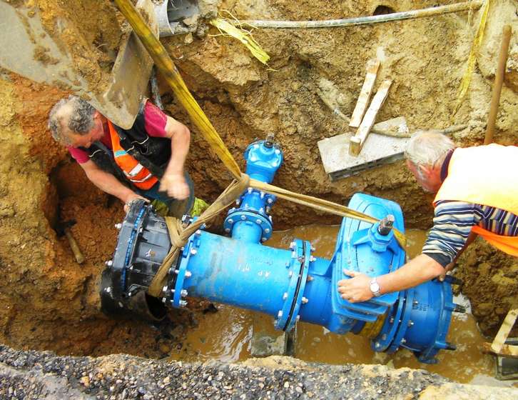 Two construction workers in high-visibility vests fitting a large blue water pipe assembly in a muddy trench
