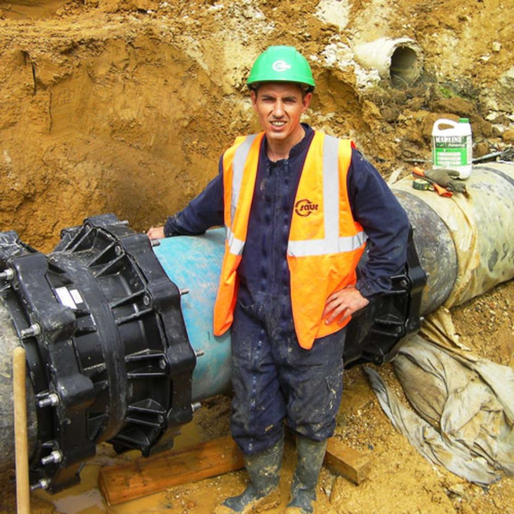 Utility worker in a high-vis vest and hard hat standing in an excavation trench beside a large pipe joint
