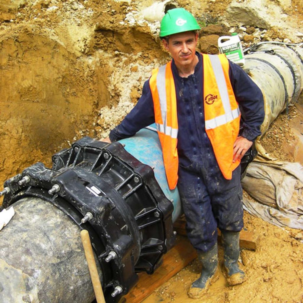 Utility worker in a green hard hat and high-vis vest standing beside a large pipe coupling in an excavation trench