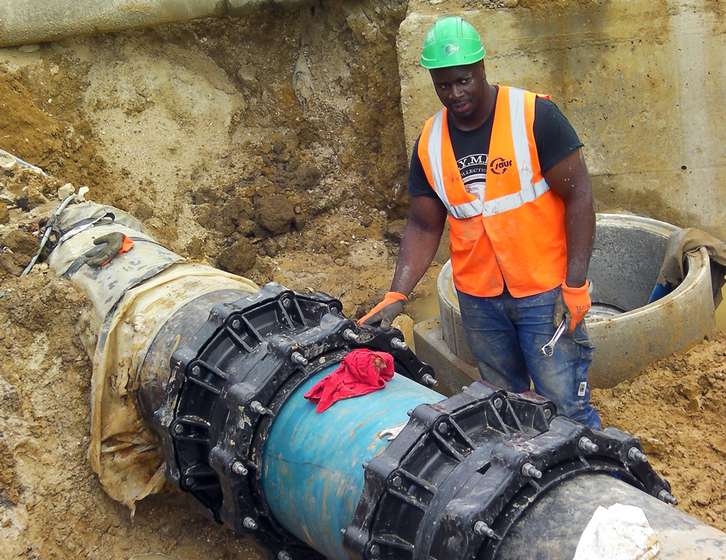 A construction worker wearing a hard hat and hi-vis vest working on a large underground pipe joint in a trench