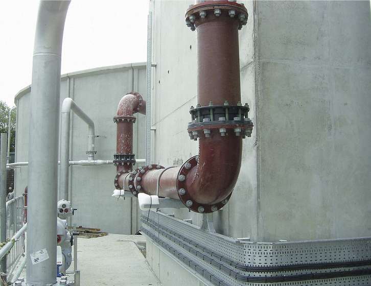 Red industrial pipework with bolted flanged joints attached to large concrete storage tanks
