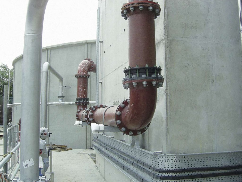 Reddish-brown industrial piping with flanged connections running along the exterior wall of a concrete storage tank