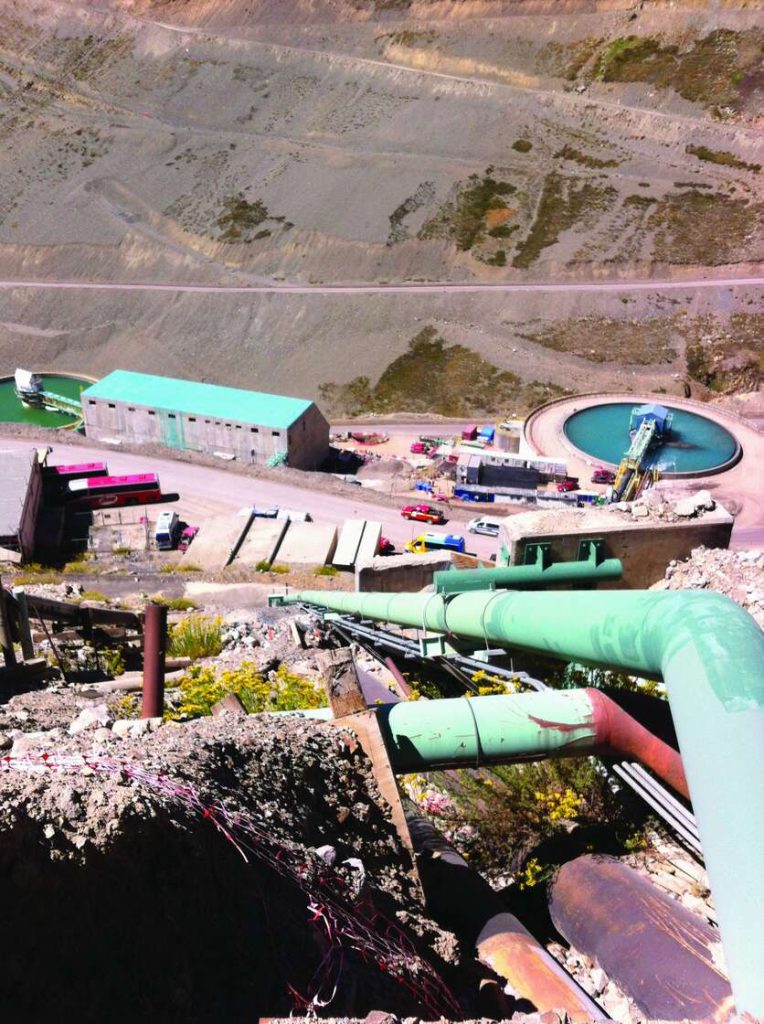 Large green pipes descending a steep mountain slope towards industrial mining buildings and circular pools