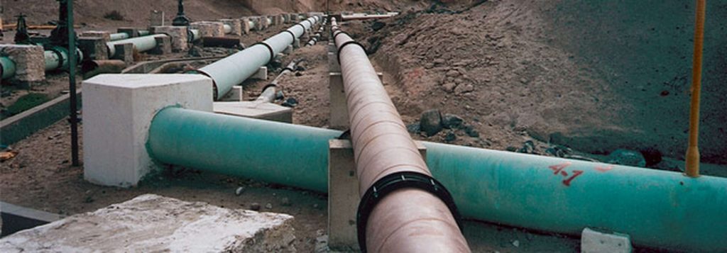 Large industrial pipelines supported on concrete blocks running across a dusty, desert terrain
