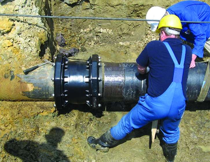 Two utility workers in hard hats fitting a mechanical coupling onto a large pipe in an excavated trench