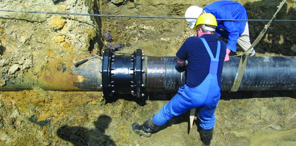 Two utility workers in hard hats installing a mechanical coupling on a large underground pipe in a trench