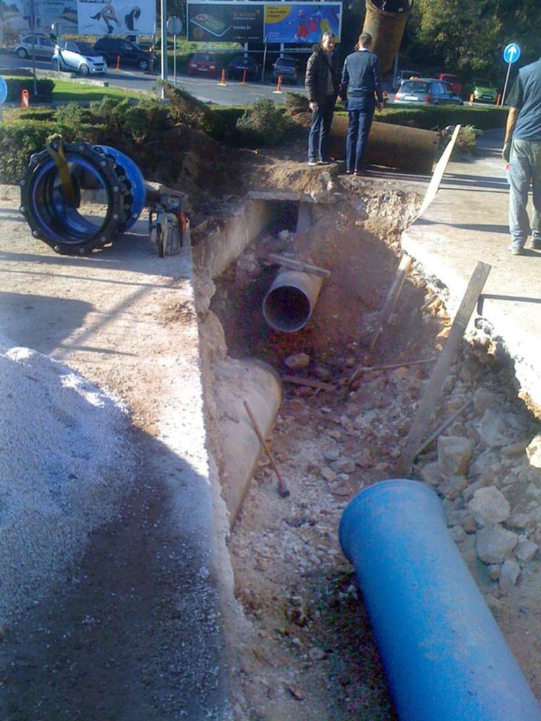 Construction workers beside a deep excavated trench exposing large underground water pipes