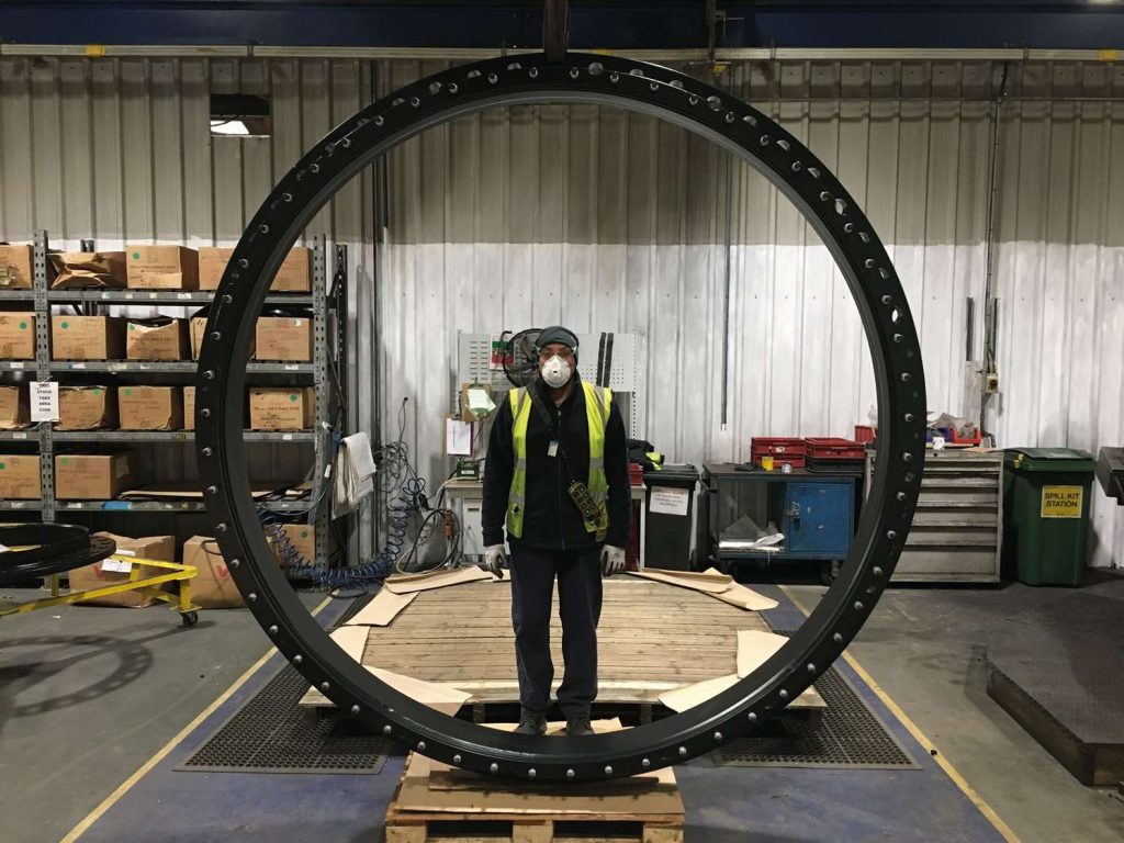 A worker in a hi-vis vest and face mask standing inside a massive black industrial metal ring on a factory floor
