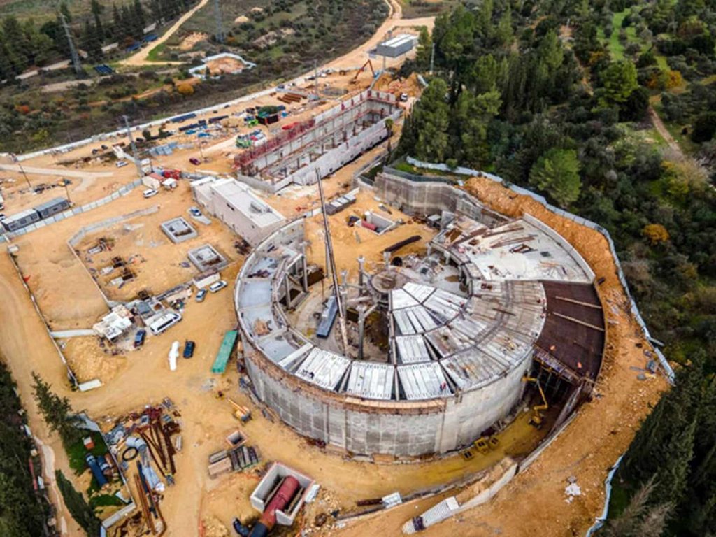 Aerial view of a large circular concrete structure under construction surrounded by construction equipment and trees