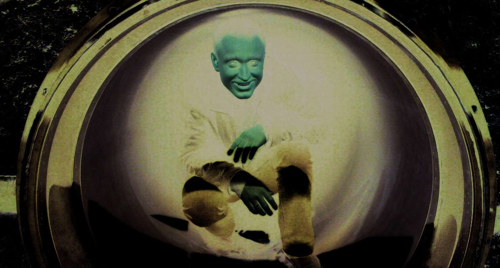 A person with green skin smiles while crouched inside a large circular hatch, shown in inverted colours