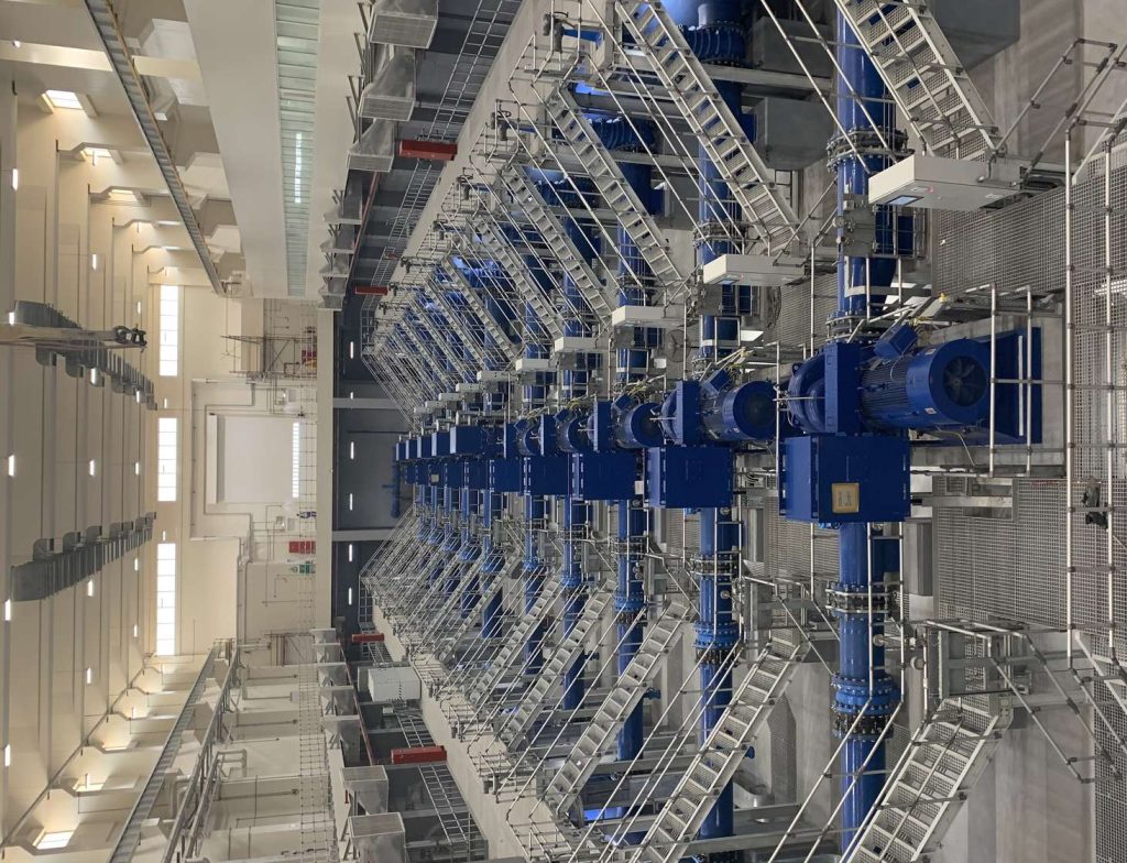 Rows of large blue pipes, pumps, and metal catwalks inside a spacious industrial facility