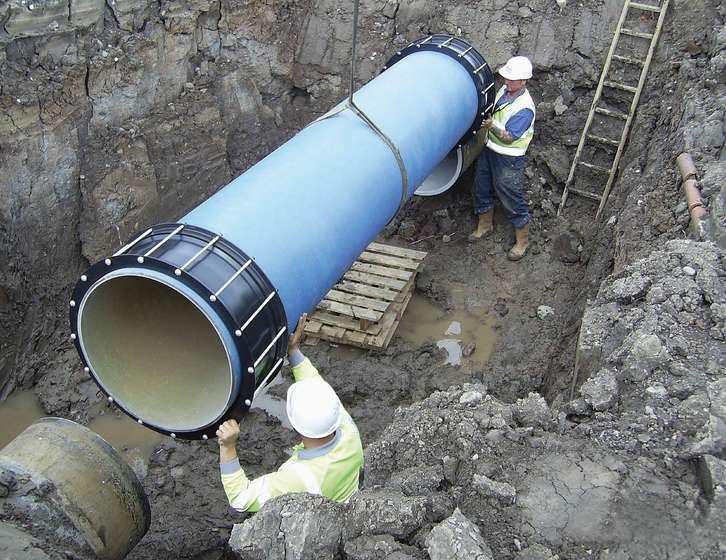 Two construction workers in high-vis clothing guide a large blue pipe into a trench