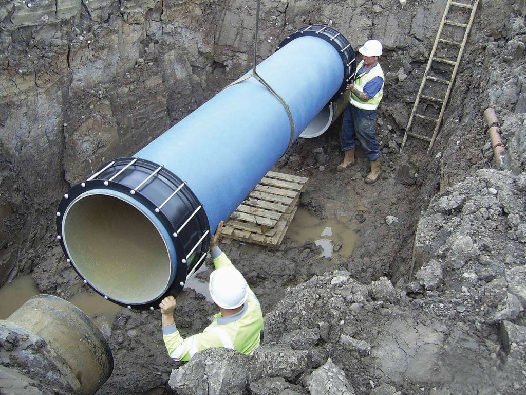 Two construction workers guiding a large blue pipe into a muddy trench
