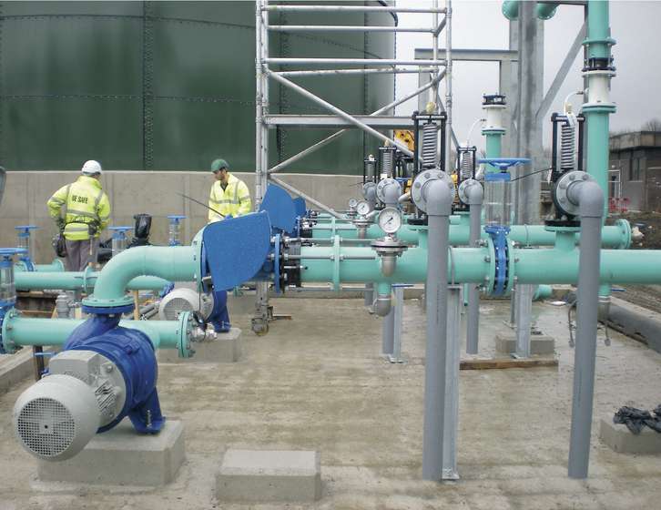 Two workers in high-visibility jackets near large industrial piping, pumps, and valves at an outdoor facility