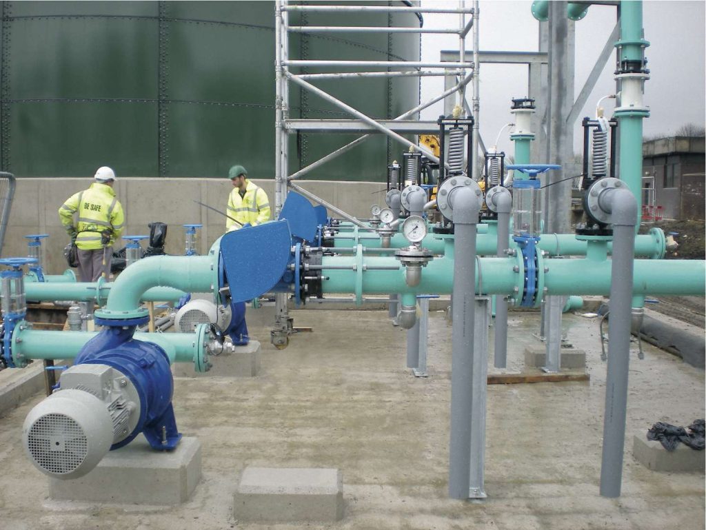 Two workers inspecting industrial pipework, blue pumps, and pressure gauges at a plant