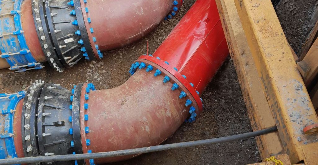 Large industrial pipes with flanged elbow joints secured with blue bolts in an excavation trench