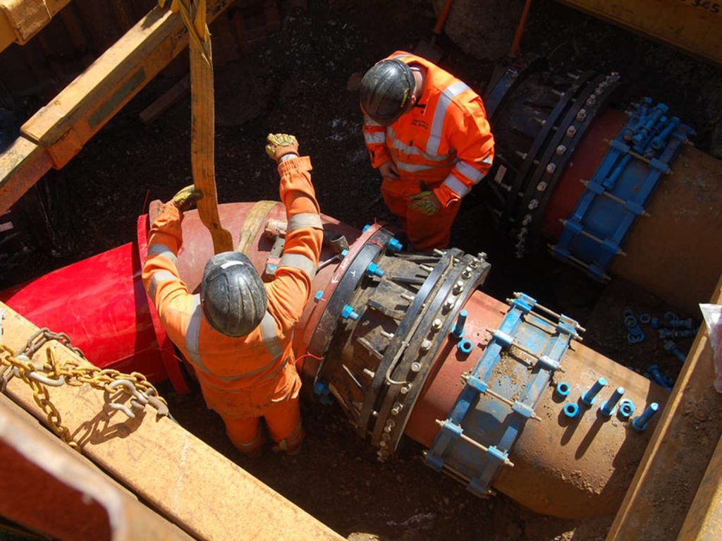 Two workers in high-visibility orange overalls and hard hats installing large underground pipework in a trench