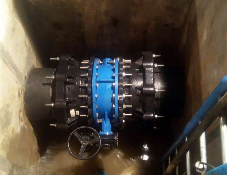 Large blue valve connected to black pipes with heavy bolts inside an underground concrete chamber