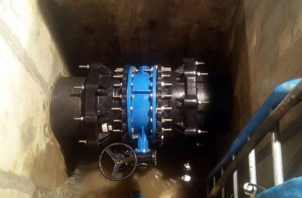 Large blue valve with a handwheel mounted on black industrial pipework inside a concrete chamber