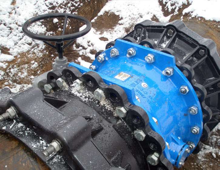 Large blue industrial pipe valve with a manual handwheel and heavy bolted couplings on snowy ground