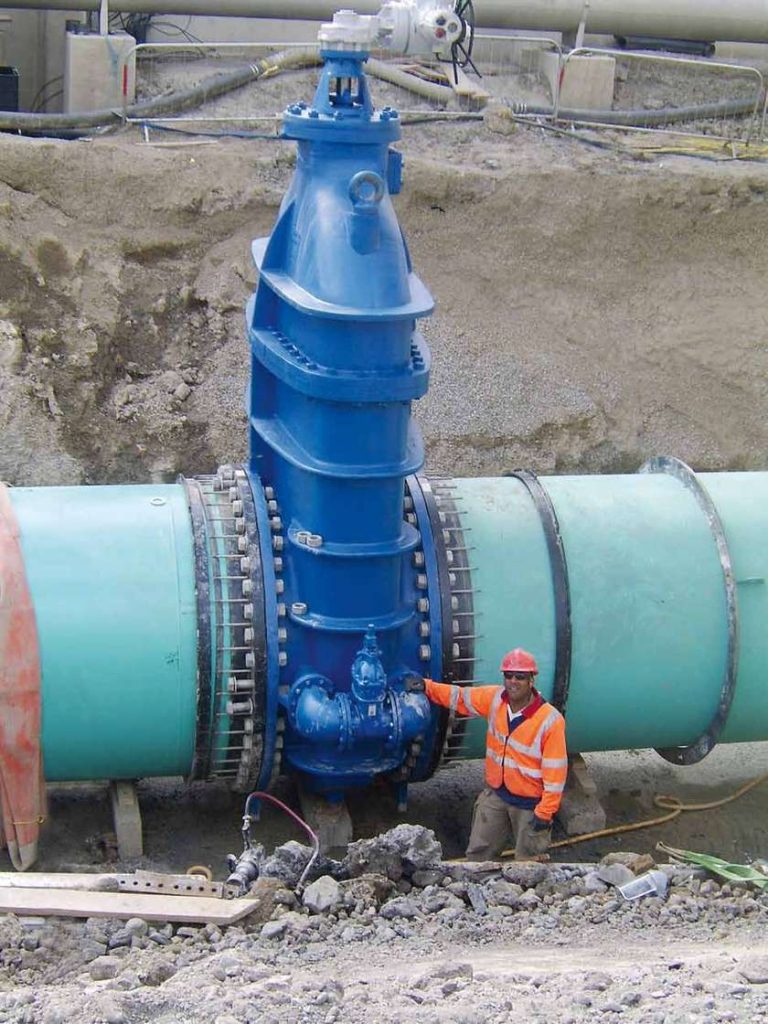 A construction worker standing next to a massive blue gate valve on a large pipeline to show scale
