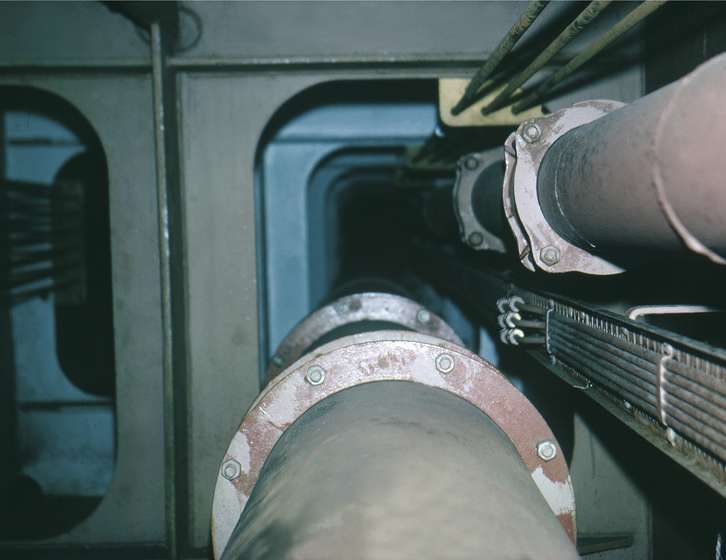 Large pipes with bolted flanged joints and cable trays running along a narrow steel bulkhead tunnel