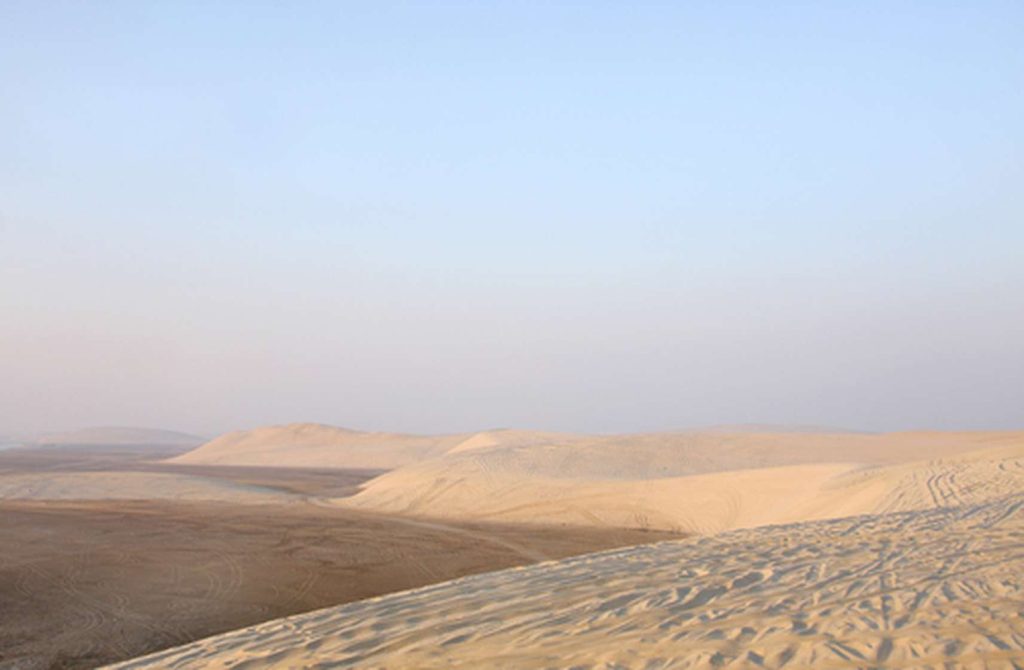 Vast desert landscape featuring undulating sand dunes with tyre tracks under a clear pale sky