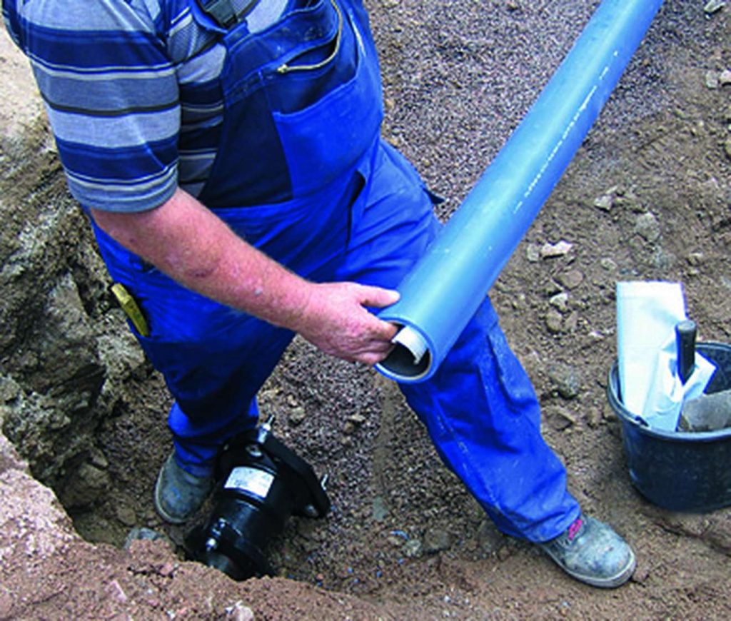A worker wearing blue dungarees holds a large blue pipe in an excavated trench