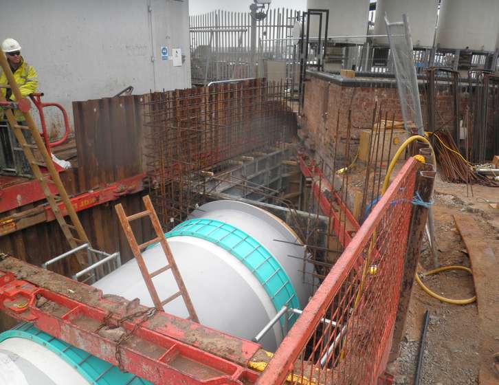 Large cylindrical pipe being installed in a shored excavation pit at a construction site